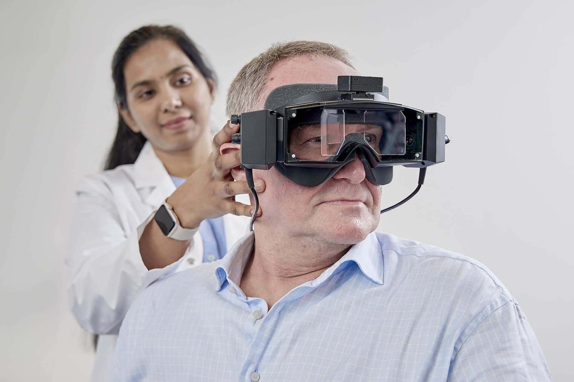 Frontal view of patient wearing VNG goggles. A clinician stood behind the patient is turning the patient's head 45 degrees to the left