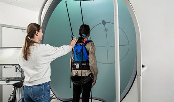 Physical therapist stood behind patient, with their right hand held up a short distance behind the patient's upper back. The patient is stood inside the Bertec CDP/IVR virtual reality dome on the dynamic platform. The patient is strapped into a harness and performing an exercise where they have to lean forward, sideway, and backward.