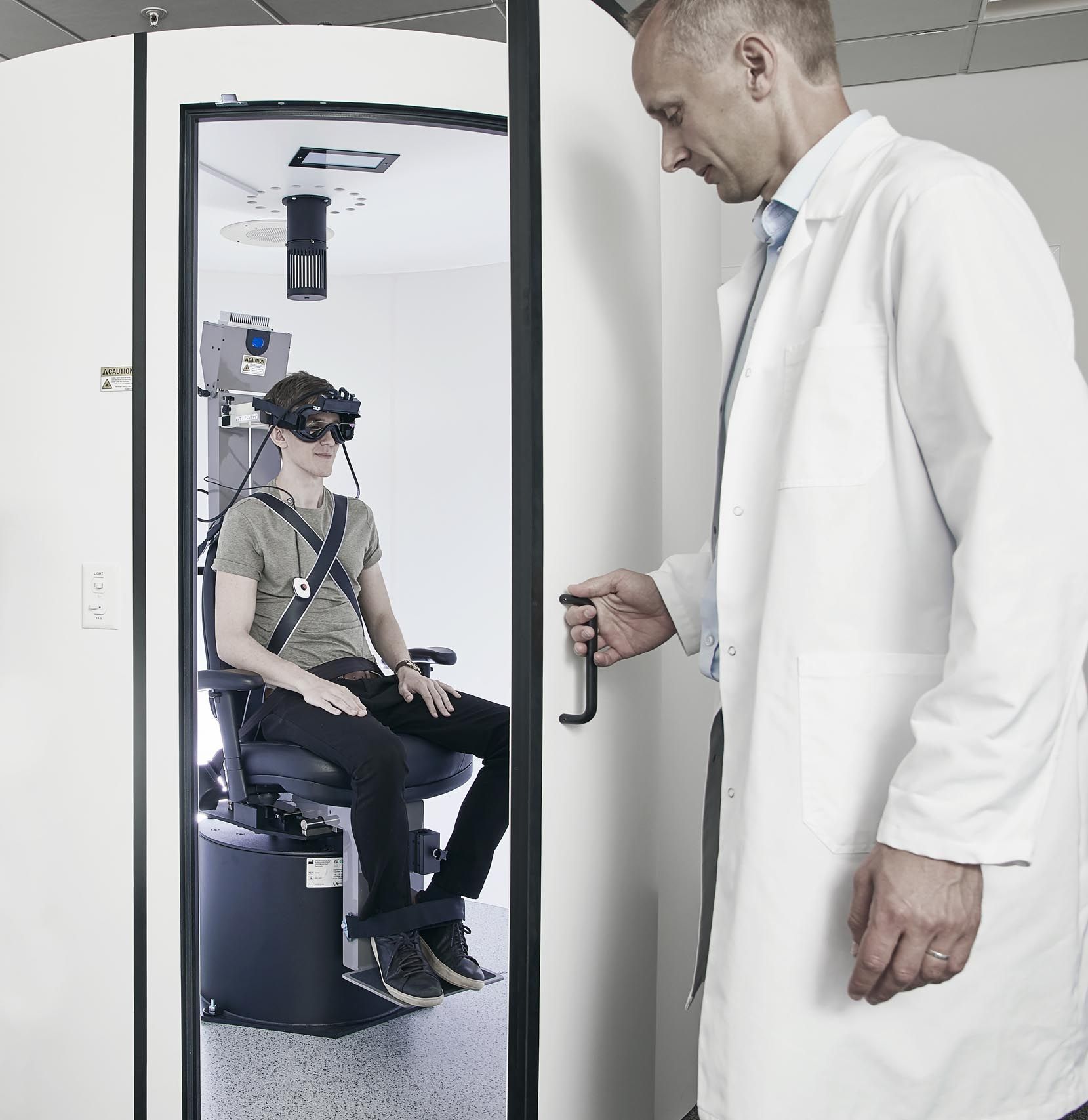 Male clinician closing booth door for Orion Auto-Traverse chair. Male patient fixated in the chair wearing VNG goggles