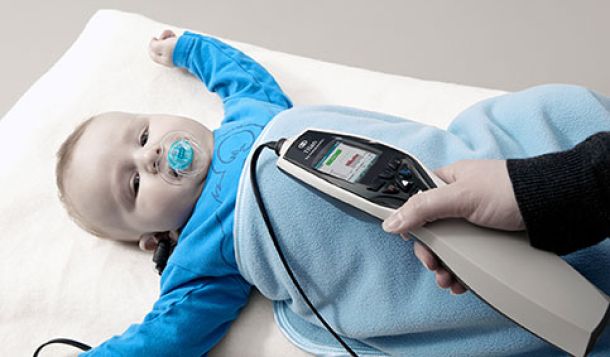 Clinician performing TEOAE in a small baby boy. The boy is on his back with his arms raised above his head. He is looking cheerful and smiling, has a blue pacifier in his mouth, is wearing a blue jumper and covered by a blue blanket.